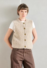 Kratka, bežna pletena brezrokavnik z teksturiranim zaključkom, z okroglo ovratnikom in petimi zelenkastimi gumbi spredaj. Nosi se čez belo majico.