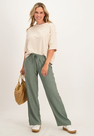 Vrouw glimlacht, draagt een beige gestreept overhemd, groene broek, witte sneakers, houdt een gevlochten beige handtas vast, staand op een effen lichte achtergrond.