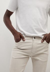 Pantalones de algodón beige claro con un corte clásico, que cuentan con bolsillos frontales, cierre con botón y una textura suave. Combinados con una camiseta blanca.