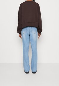 Osoba w luźnej, ciemnobrązowej bluzie i jasnoniebieskich dżinsach o prostym kroju, stojąca przodem do białej ściany z relaksującymi rękami.