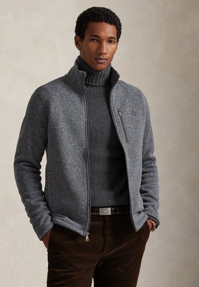 Chaqueta gris de forro polar con cremallera y cuello alto, sobre un suéter de punto a juego, combinada con pantalones de pana marrones. Incluye detalle de bolsillo lateral con cremallera.