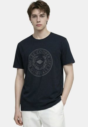 Jeune homme aux cheveux brun foncé portant un t-shirt noir Lee Cooper avec un logo circulaire, associé à un pantalon blanc, debout devant un fond uni.