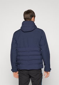 Σκούρο μπλε μπουφάν με κουκούλα, με καπιτονέ σχέδιο στο κάτω μέρος της πλάτης και κόντρα λείο ύφασμα στην πάνω περιοχή.