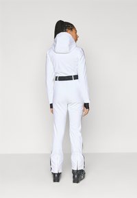 Λευκή στολή σκι με κουκούλα, μαύρες λεπτομέρειες σε μανσέτες και πλάγια, εφαρμοστή σχεδίαση, μαύρη ζώνη με λογότυπο στη μέση, φορεμένη με μαύρα μπότς σκι.