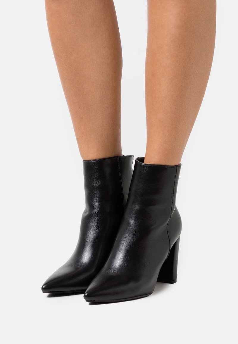 Bottes bottines en cuir noir avec un bout pointu et un talon bloc. Texture lisse, design minimaliste, et ajustement près de la cheville.