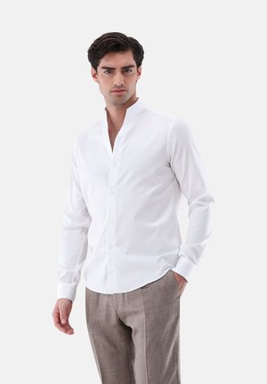 Camicia bianca a maniche lunghe con colletto alla cinese, chiusura con bottoni, tessuto liscio, stile aderente, indossata con pantaloni a quadri color marrone chiaro.