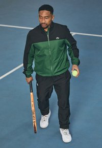 Sportiv îmbrăcat într-o jachetă verde și neagră, cu fermoar, și pantaloni negri, ținând o rachetă de tenis și o minge, purtând adidași albi.