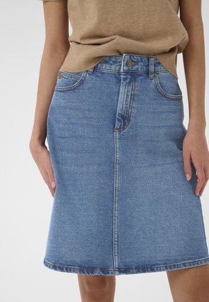 Jupe en jean bleu mi-longue avec couture frontale, bouton et poches, portée avec un haut beige par une personne debout devant un fond uni.