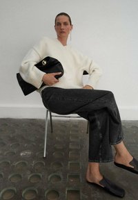 Suéter crema oversize, clutch negro, jeans grises. Textura suave, corte relajado y zapatos negros sin cordones. Estilo minimalista sobre una silla.