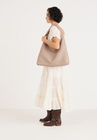Beige strukturierte Tragetasche mit einem weichen, lässigen Design, über der Schulter getragen. Die Modells trägt ein weißes, gestuftes Kleid und braune kniehohe Stiefel.