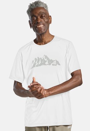 Uomo anziano sorridente con capelli grigi che indossa una t-shirt bianca con grafica montana e pantaloni beige, con le mani incrociate davanti.