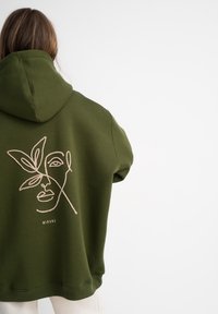 Haut à capuche vert olive avec un grand motif brodé représentant un visage et une feuille au dos, offrant une coupe décontractée et une texture douce.