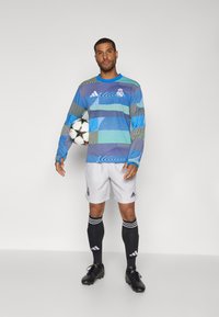 Langarm blauw-groen gemusterde jersey met ronde hals, gecombineerd met grijze shorts en zwarte sokken met witte strepen. Zwarte voetbalschoenen.