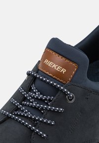 Chaussure à lacets bleu marine avec des accents en cuir, présentant un tissu texturé, des lignes blanches contrastantes sur les lacets et un patch en cuir tan avec logo.