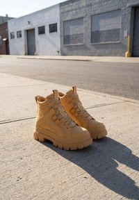Paire de grosses bottines camel à lacets sur un trottoir en béton avec un bâtiment industriel et une rue en arrière-plan.