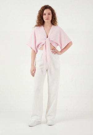 Femeie în picioare, purtând o bluză roz deschis cu mâneci scurte legată, pantaloni albi largi și adidași albi, în fața unui fundal alb simplu.