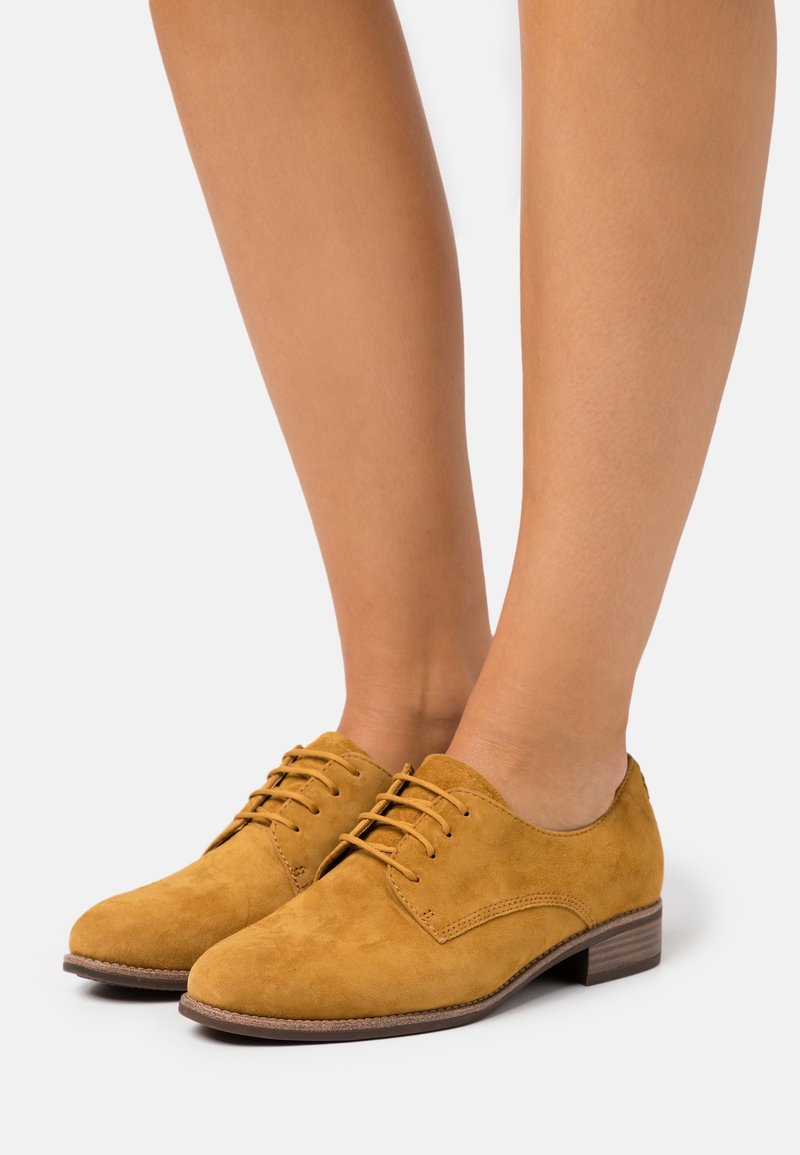 Paire de chaussures Oxford lacées en daim jaune moutarde portées pieds nus sur un fond uni.