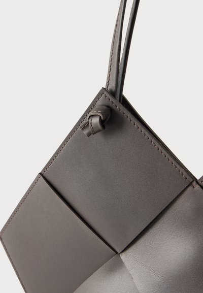 Sac à main en cuir gris foncé avec coutures visibles, détail de sangle nouée et design à panneaux géométriques sur fond blanc uni.