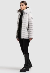 Světlounce šedá prošívaná bunda s vysokým límcem, horizontálním prošíváním, dvěma postranními zipy a černou kovovou výbavou, doplněná o černé legíny a kozačky po kolena.