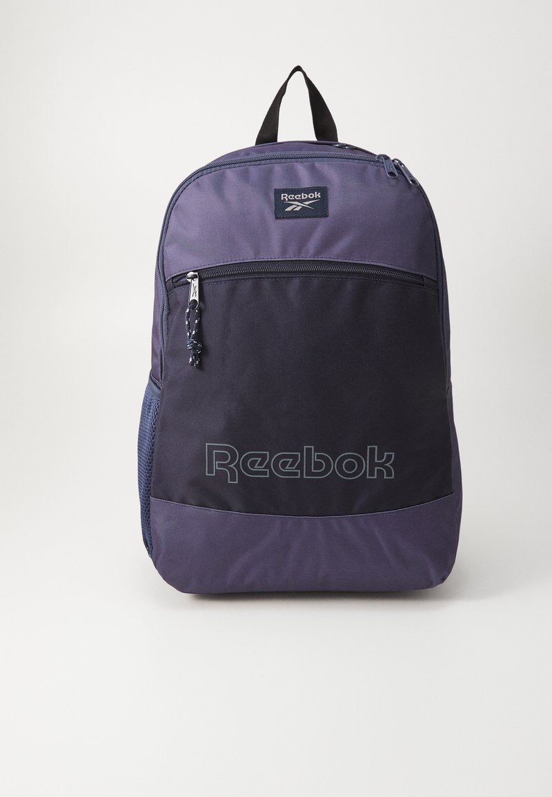 Reebok kuprinė tamsiai violetinės ir juodos spalvos audinyje, su viršutiniu rankenom, priekinėmis užtrauktukinėmis kišenėmis ir logotipu apatinėje priekinėje dalyje. Lengvas dizainas.