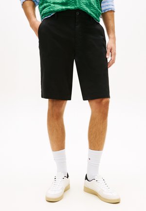 Mand iført sorte knælange shorts, hvide ankelsokker, hvide sneakers, med hænderne i lommerne, stående på en ensfarvet baggrund.