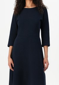 Tmavě modré šaty s dlouhým rukávem a kulatým výstřihem, texturovaná látka, přiléhavý živůtek a rozšířená sukně, vystihující čisté linie a minimální zdobení.