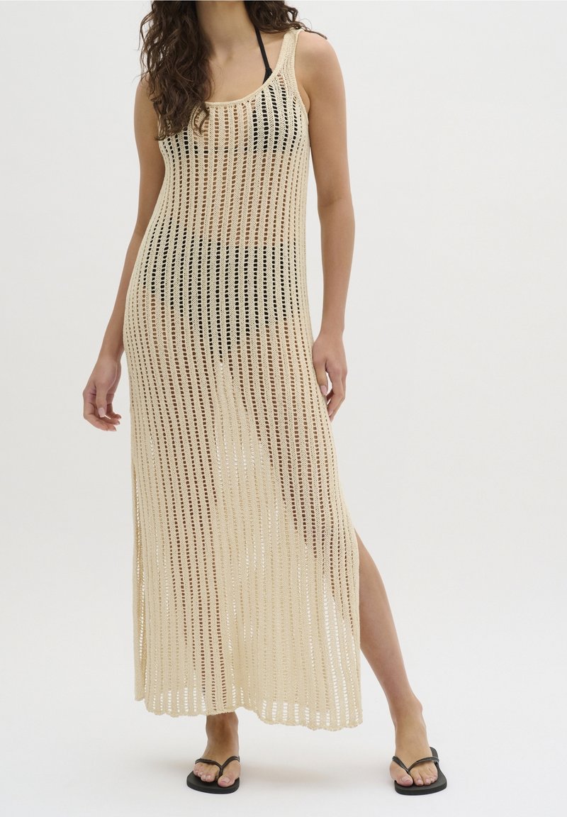 Robe par-dessus en maille beige à rayures verticales, avec un col rond et des fentes latérales, portée avec des tongs noires.