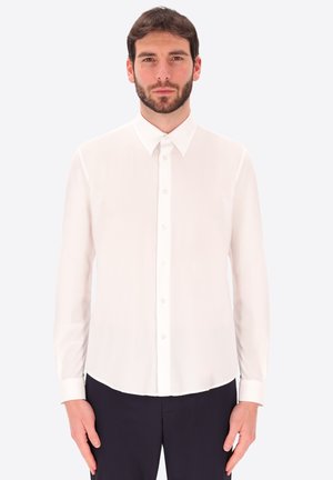 Uomo con capelli scuri e barba indossa una camicia bianca abbottonata e pantaloni neri, in piedi davanti a uno sfondo chiaro e uniforme.
