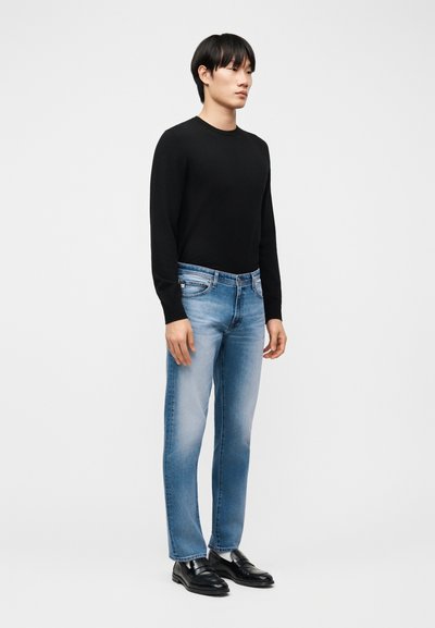 Νέος άνδρας που στέκεται φορώντας μαύρο μακρυμάνικο πουκάμισο, ανοιχτόχρωμο μπλε τζιν, λευκές κάλτσες και μαύρα δερμάτινα μοκασίνια σε απλό φόντο.