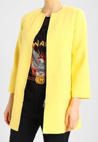 Chaqueta amarilla con cremallera, hecha de una tela suave, que presenta un cuello redondo, bolsillos laterales y mangas de tres cuartos sobre una camiseta gráfica.