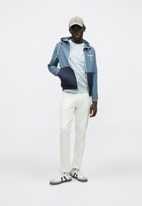 Ζακέτα με φερμουάρ σε μπλε και ναυτικό μπλοκ χρωμάτων με λευκές λεπτομέρειες, ανοιχτό μπλε μπλουζάκι, άσπρο παντελόνι και αθλητικά γκρίζα παπούτσια με μαύρες ρίγες.