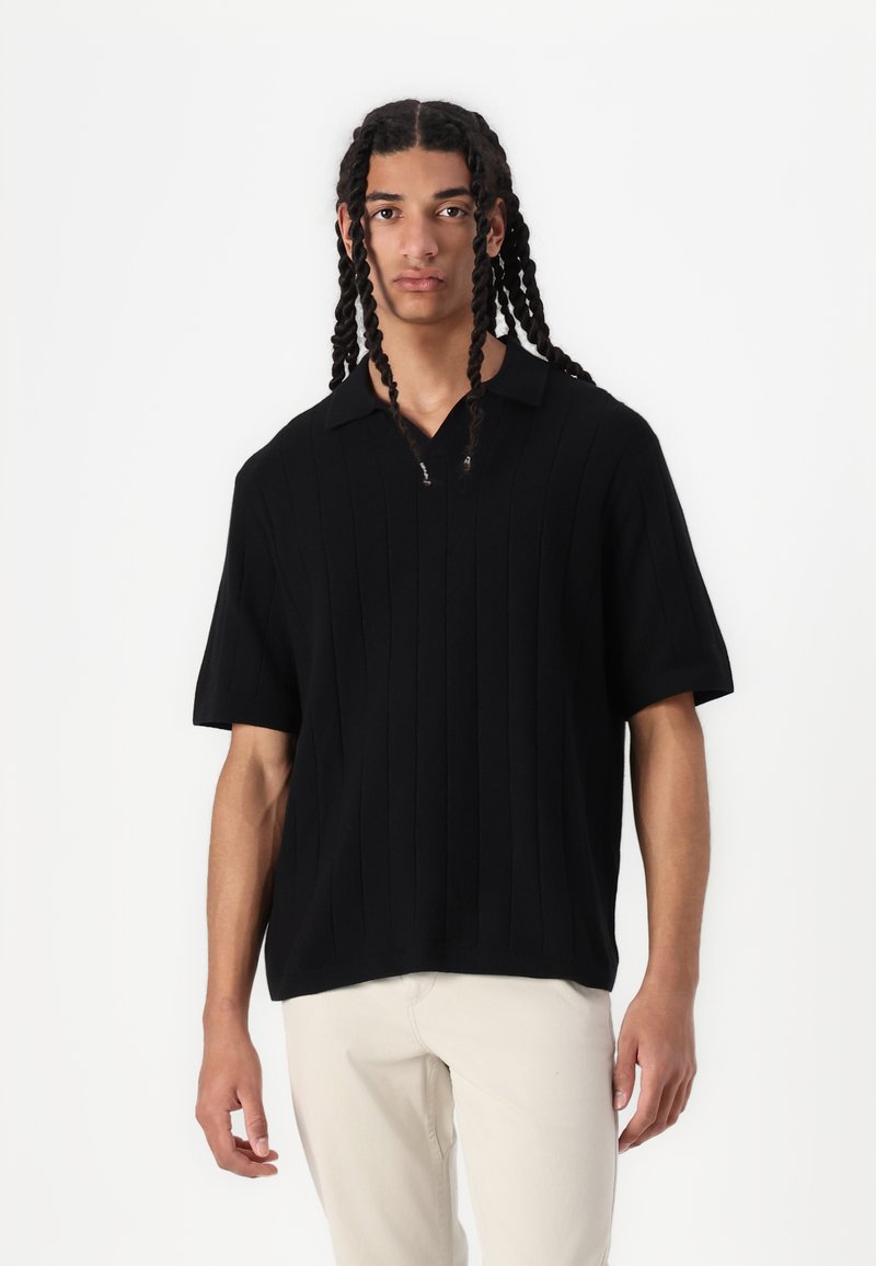 Černé polo triko s krátkým rukávem z pleteniny s ribbed texturou, véčkovým výstřihem a zipovým detaily, spárované se světlými kalhotami.