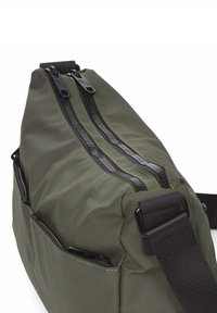 Bolsa de tela verde oliva con múltiples cremalleras negras, costuras amarillas en contraste y una correa negra ajustable. Cuenta con varios bolsillos exteriores.