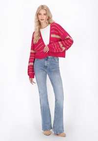 Felle roze cropped cardigan met gouden en witte strepen, gecombineerd met lichtblauwe flared jeans. Textuur gebreide stof, casual stijl.