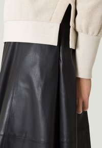 Beige gebreide top met een brede manchet, gecombineerd met een zwarte leren rok met zichtbare naden en een gladde, glanzende textuur.