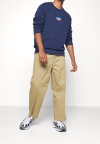 Person som bär en marinblå Levi's sweatshirt, beige byxor och vit-grå sneakers som står mot en vit bakgrund.