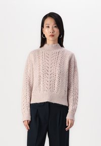 Világos rózsaszín kötött pulóver magas nyakkal, textúrált mintázattal és nyitott öltésekkel, sötét nadrággal párosítva.
