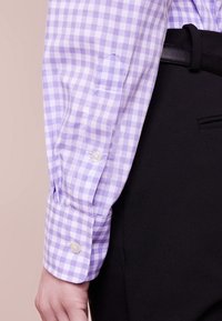 Lila och vit rutmönstrad långärmad skjorta med knäppta manschetter, tillsammans med svarta byxor och ett svart bälte, synlig på en persons arm och midja.