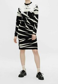 Robe tricotée avec un motif audacieux en zigzag noir et blanc, dotée d'un grand col en dentelle blanche et de manches longues, assortie à des chaussures noires.
