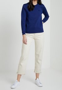 Laipi su jūrų jėgos puloveriu, su apvaliu kaklu ir įsiūtomis rankovėmis, suderinta su baltais džinsais su plaukuota apatine dalimi ir baltais sportbačiais. Minimalistinis dizainas.