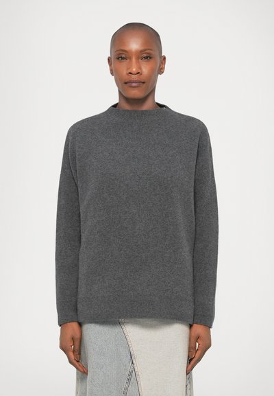 Femme avec la tête rasée portant un pull gris foncé et une jupe en denim claire avec une couture avant, se tenant contre un fond blanc uni.