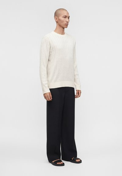 Άνδρας που φοράει λευκό πλεκτό πουλόβερ με σχέδιο κοτσίδας, μαύρο φαρδύ παντελόνι και μαύρα σανδάλια, στέκεται μπροστά σε απλό γκρι φόντο.