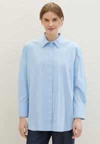 Femme portant une chemise à boutons légère, bleu clair, à col et à manches longues, associée à un pantalon foncé, sur un fond uni.