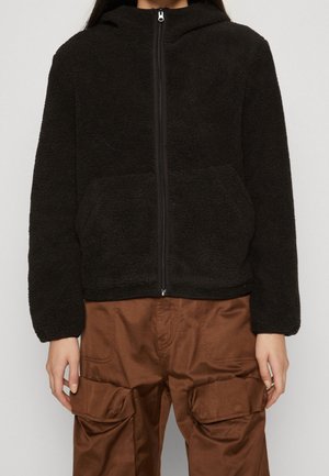 Person wearing a black zip-up fleece jacket and brown cargo pants with large front pockets, standing against a plain background.