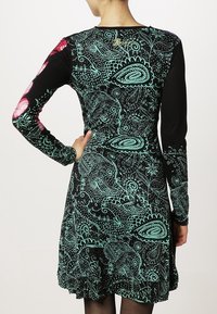 Nainen, joka wear musta pitkähihainen mekko, jossa on turkoosia paisley- ja kukkakuvioita, sekä vaaleanpunaisia kukkakirjailuja yhdessä hihassa, kääntyy poispäin.