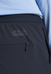 Detailní záběr na černé kalhoty Jack Wolfskin s kapsou na zip a síťovaným páskem, nošené s bledě modrým topem.