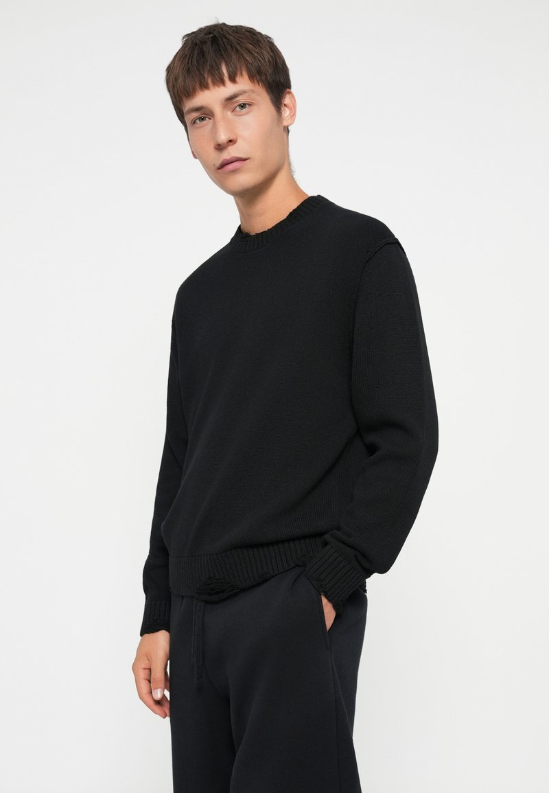 Zwarte gebreide crew neck trui met geribde manchetten en onderkant, gecombineerd met zwarte pantalons. De textuur lijkt glad en iets losser passend.