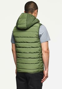 Grüne gepolsterte Weste mit Kapuze, versehen mit einem gesteppten Design, strukturiertem Stoff und Seitennähten, getragen über einem grauen Hemd.