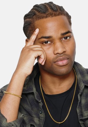 Portrait d'une personne aux cheveux tressés, portant une chemise sombre et une veste à carreaux verte. Les détails notables incluent une chaîne en or et des bracelets comme accessoires.