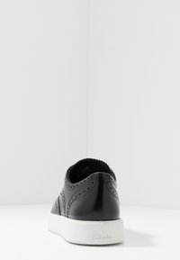 Chaussure en cuir noir avec design perforé et bout arrondi. Semelle coussinée et semelle extérieure en caoutchouc blanche contrastée. Nom de la marque affiché sur le côté.